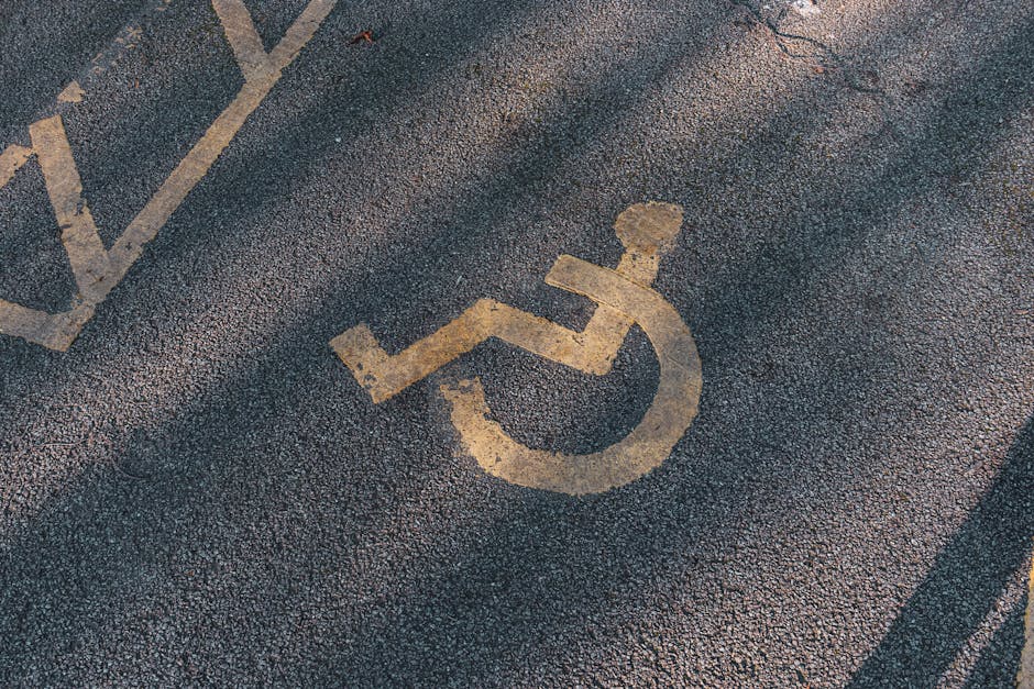 A close-up view of a designated accessible parking space on a rough asphalt surface, featuring a faded yellow wheelchair symbol painted on the ground. The symbol, which depicts a person seated in a wheelchair, is slightly worn but clearly visible. Surrounding the symbol are faint markings for nearby parking bays, including a partially visible rectangular box and lines indicating parking boundaries. The area is likely part of a residential or commercial parking lot adjacent to a property, possibly outside a home or building undergoing a house removal or relocation process. This parking space may be used for loading and unloading furniture and boxes during the home relocation, with the pavement and markings maintained for vehicle access and convenience. The image context aligns with professional removals services such as those provided by Removals Enfield, which assist with packing, loading, and transport logistics, especially around enfield properties on Southbury Road.