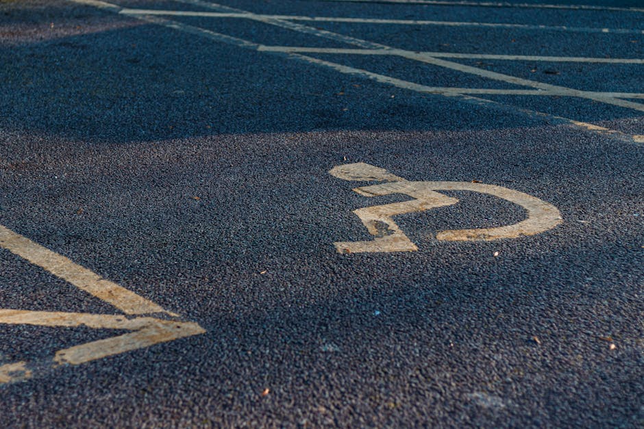 Close-up view of a yellow painted wheelchair accessible parking symbol on an asphalt surface, with visible tire marks and parking bay lines in the background. The parking area appears to be outdoors, with natural lighting and no vehicles present. This image illustrates designated disabled parking spaces commonly found near residential or commercial properties, such as homes or offices in Southbury Road, Enfield. It reflects the importance of accessible parking during house removals or domestic moves, supported by companies like Removals Enfield, which specialise in home relocation and furniture transport. The overall scene emphasizes the infrastructure necessary for smooth logistics during packing and loading activities, with clear markings to facilitate appropriate vehicle and equipment placement for efficient and accessible furniture transport.