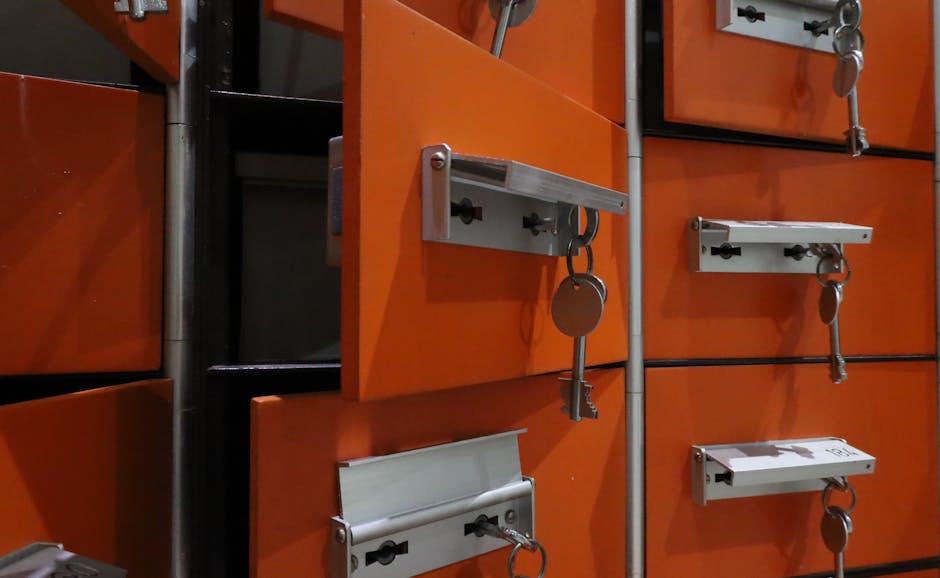A close-up view of orange locking cabinets used for secure storage, with metal locking bars and keys hanging from small key hooks attached to each lock. The cabinets are located inside a property during a home relocation process, with some doors slightly open, revealing a glimpse of dark interior compartments. The scene includes a surrounding metal framework and a concrete or tiled floor, indicating an organized area for storing valuable or heavy items. The locks and keys are part of the packing and moving logistics managed by Removals Enfield, involved in a house removals or furniture transport service, capturing the careful handling and securing of belongings during the loading process on a moving day.