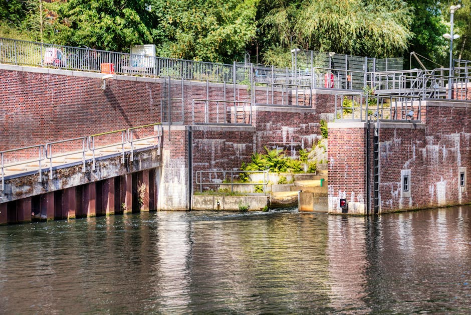 A brick canal lock with metal railings and stairs leading down to the water's edge, situated beside a body of water with calm reflections. Green vegetation and trees are visible behind the lock structure, which includes several small access points and flood gates. The area is well-lit through natural daylight, showing signs of maintenance and minor weathering on the brickwork and metal surfaces. This scene depicts the exterior environment during a home relocation process involving heavy item removals, with the canal access suggesting a logistical route for transporting large furniture or appliances from the property. The image provides a clear view of the structural features relevant to moving logistics managed by Removals Enfield, emphasizing the practical aspects of loading and transport in an outdoor setting near water.