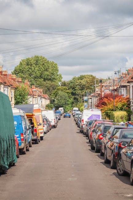 A residential street in Enfield with closely spaced terraced houses on either side. The street is lined with parked cars of various makes, models, and colours, occupying both sides of the narrow asphalt road. Several moving vans and boxes wrapped in plastic and cardboard are visible, indicating a home relocation process. Some furniture, such as a wooden chair and cardboard boxes filled with household items, are positioned near the entrance of a house. A large green tree and smaller shrubs add greenery to the scene, with an overcast sky overhead and power lines stretching across the street. The image captured during daylight showcases the typical environment for house removals in Enfield, highlighting the challenges of navigating narrow streets and terraced homes during furniture transport and loading activities, with a focus on careful handling by professionals like Removals Enfield.