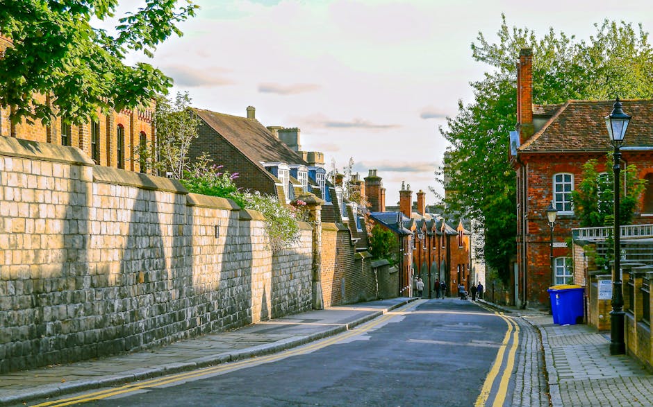 A quiet residential street in Enfield with narrow pavement and a gently curving tarmac road marked by double yellow lines. On the left, a high stone wall runs along the sidewalk, partially shading the pavement with shadows cast by overhanging branches from nearby trees. Behind the wall, rows of traditional terraced houses with brick facades, slate roofs, and chimneys are visible under the afternoon light, some with small front gardens or plants. On the right side, a red-brick building features white-framed windows and a black vintage-style street lamp attached to the facade, alongside a blue recycling bin placed on the sidewalk. Several people are seen in the distance, walking along the street, which is relatively quiet. This setting exemplifies typical house relocation and furniture transport challenges within historic terraced homes, emphasizing the importance of professional removals services like those offered by Removals Enfield for packing, loading, and transport in urban areas such as this.