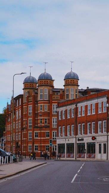 A multi-story red-brick building with distinctive rounded towers topped by gray domed roofs, situated at a street corner under a partly cloudy sky. The building features numerous white-framed windows 
